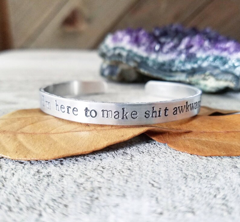 May include: A silver cuff bracelet with the stamped phrase "I'm here to make shit awkward." The bracelet is resting on a textured surface with a dried leaf and a purple crystal in the background. The bracelet is a simple, yet bold accessory.