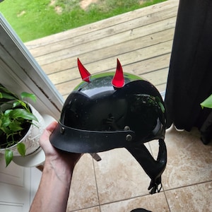 May include: A black motorcycle helmet with red devil horns attached to the top. The helmet has a black leather strap and a black visor.