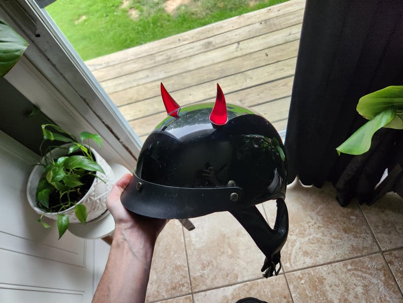 May include: A black motorcycle helmet with red devil horns attached to the top. The helmet has a black leather strap and a black visor.