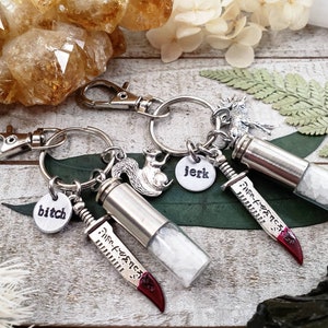 May include: Two silver keychains with small vials filled with white crystals. Each keychain has a charm, one with a squirrel, the other with a moose. The vials have a small knife charm attached with the words "Mis-Fortune" inscribed on the blade. One charm has the word "bitch" on it, the other has the word "jerk" on it.