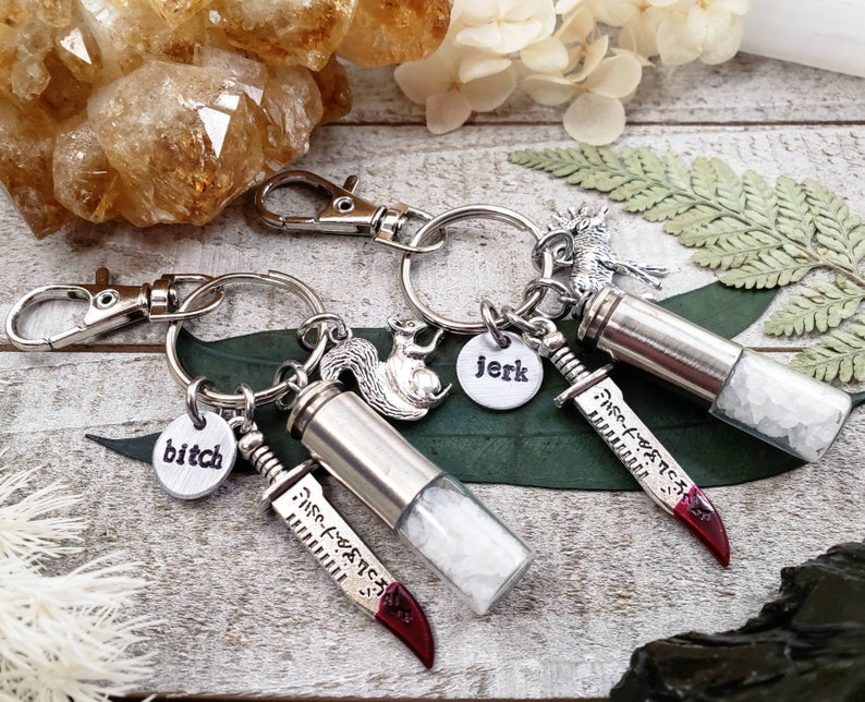 May include: Two silver keychains with small vials filled with white crystals. Each keychain has a charm, one with a squirrel, the other with a moose. The vials have a small knife charm attached with the words "Mis-Fortune" inscribed on the blade. One charm has the word "bitch" on it, the other has the word "jerk" on it.