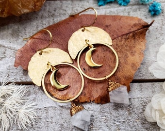 Peut inclure: Boucles d'oreilles en forme de croissant de lune dorées avec un pendentif en cristal de quartz transparent suspendu à chaque cercle. Les boucles d'oreilles sont présentées sur une feuille brune.