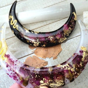 May include: Two crescent moon-shaped resin ornaments with dried flowers and gold leaf accents. One is black and the other is white.