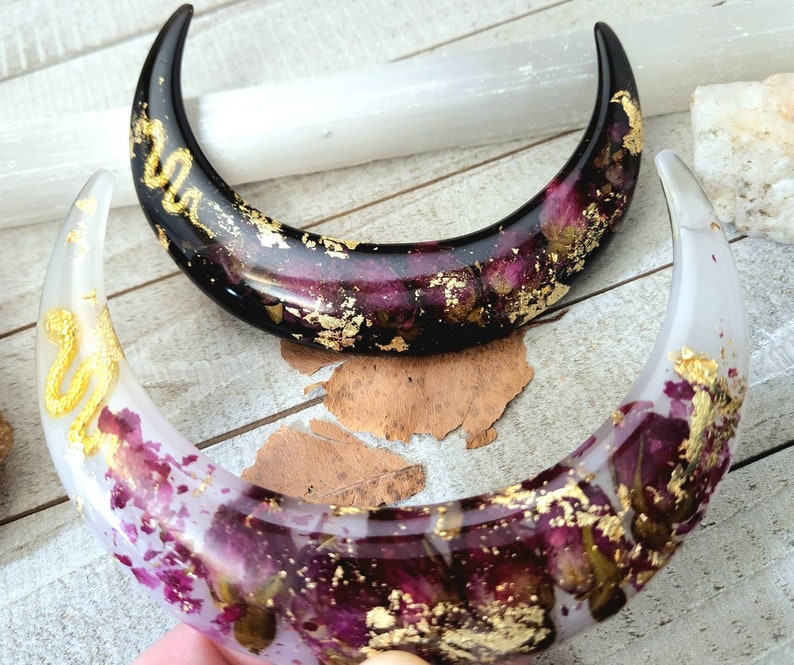 May include: Two crescent moon-shaped resin ornaments with dried flowers and gold leaf accents. One is black and the other is white.