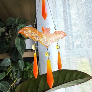 May include: A white and orange bat-shaped wind chime with crystal accents. The bat is decorated with glitter and hangs from a silver chain.