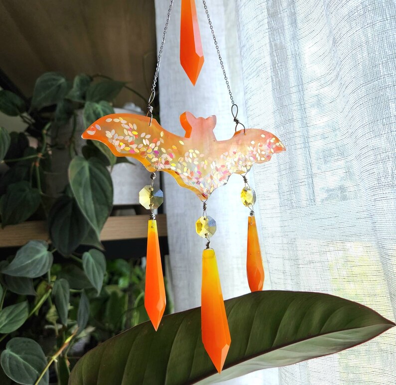 May include: A white and orange bat-shaped wind chime with crystal accents. The bat is decorated with glitter and hangs from a silver chain.