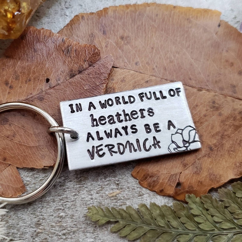 Pu&ograve; includere: Un portachiavi d'argento con un'etichetta rettangolare su cui &egrave; scritto "In a world full of heathers always be a Veronica". L'etichetta ha un piccolo disegno di fiore sul lato destro.