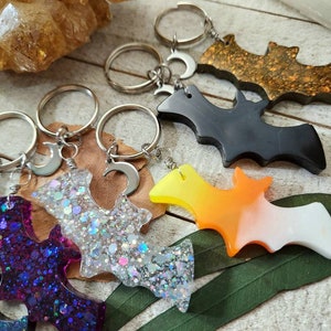 Pode incluir: Cinco chaveiros diferentes em forma de morcego com argolas de chave prateadas e pingentes de lua crescente. Os morcegos s&atilde;o feitos de resina e t&ecirc;m cores e padr&otilde;es diferentes, incluindo glitter roxo, glitter prata, preto, degrad&ecirc; laranja e branco, e glitter dourado.