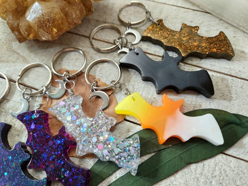 Pode incluir: Cinco chaveiros diferentes em forma de morcego com argolas de chave prateadas e pingentes de lua crescente. Os morcegos s&atilde;o feitos de resina e t&ecirc;m cores e padr&otilde;es diferentes, incluindo glitter roxo, glitter prata, preto, degrad&ecirc; laranja e branco, e glitter dourado.
