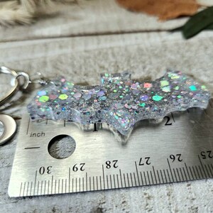 Pode incluir: Um amuleto de morcego de glitter prateado com um amuleto de lua crescente prateado preso a um chaveiro prateado. O amuleto de morcego mede aproximadamente 5 cm de largura e &eacute; medido com uma r&eacute;gua.