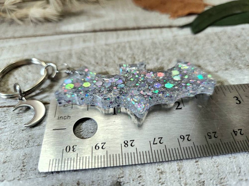 Pode incluir: Um amuleto de morcego de glitter prateado com um amuleto de lua crescente prateado preso a um chaveiro prateado. O amuleto de morcego mede aproximadamente 5 cm de largura e &eacute; medido com uma r&eacute;gua.