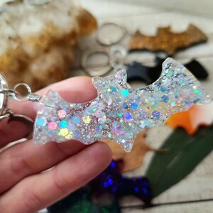 Pode incluir: Um chaveiro prateado com um amuleto de morcego de glitter branco. O morcego &eacute; feito de resina e tem glitter iridescente embutido.