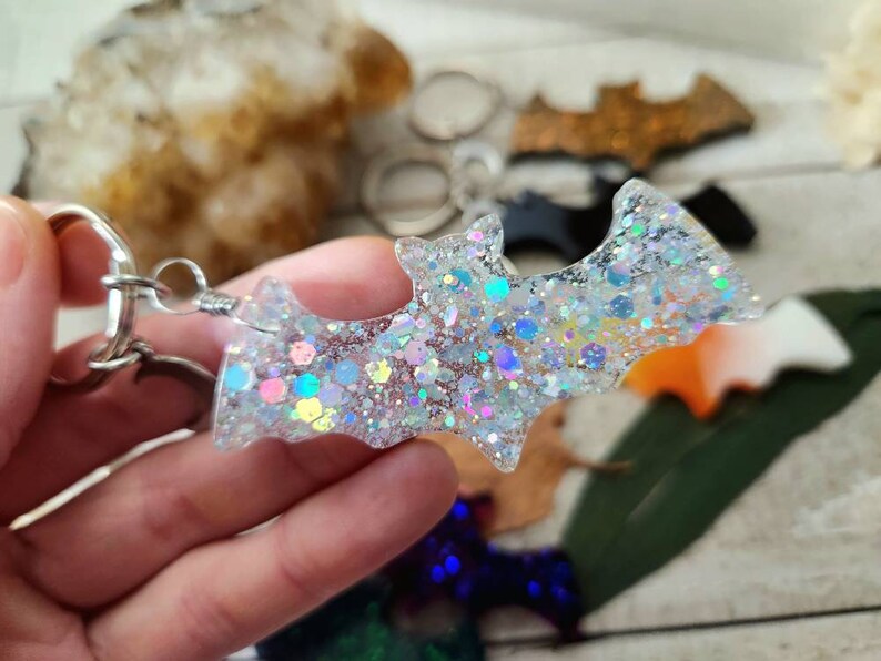 Pode incluir: Um chaveiro prateado com um amuleto de morcego de glitter branco. O morcego &eacute; feito de resina e tem glitter iridescente embutido.