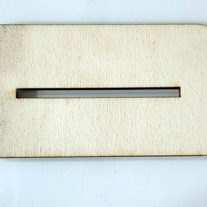 May include: A rectangular wooden craft piece with rounded corners and a long, narrow slot cut through the center. The wood has a light, natural color and visible grain. The slot appears to be approximately 0.25 inches wide.