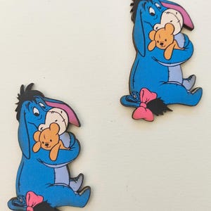 May include: Two blue Eeyore character cutouts holding a small orange teddy bear. Each cutout has a pink bow and a black mane. The background is a plain white surface.