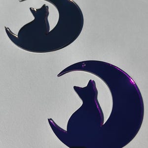 May include: Two purple cat and moon shaped ornaments. Each ornament features a cat silhouette sitting on a crescent moon. The ornaments have a reflective, metallic finish and are set against a white background.