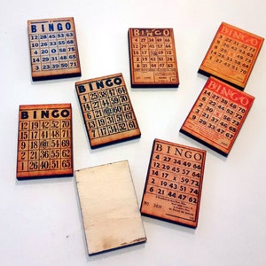 May include: Seven wooden bingo cards with black numbers and the word "BINGO" printed in a red banner. The cards are on a white surface.