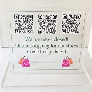 Pode incluir: Uma placa retangular de acrílico transparente com três códigos QR em preto e branco no topo. Abaixo dos códigos, o texto diz: We are never closed! Online shopping for our stores. Come in any time :). Dois ícones de sacolas de compras rosa e laranja estão na parte inferior.