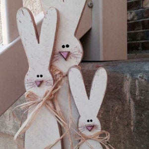 Può includere: Tre figurine di coniglio di legno bianco con nasi rosa e occhi neri. I conigli sono disposti in fila, con il coniglio più grande dietro e il coniglio più piccolo davanti. I conigli sono decorati con rafia marrone.
