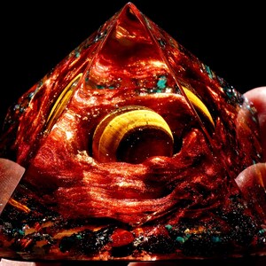 May include: A red, gold, and blue pyramid-shaped crystal with a swirling pattern inside. The crystal is made of resin and has a shiny, glossy finish.