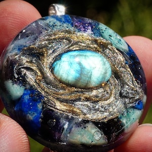 Pode incluir: Um pingente redondo de resina azul, roxo e dourado com uma grande pedra oval de labradorita azul iridescente no centro. O pingente é engastado em uma argola de metal prateado.