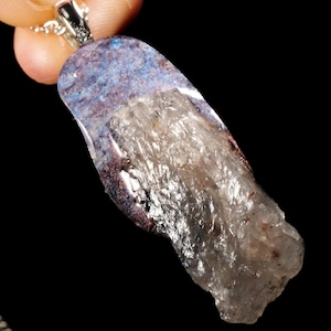 May include: A natural stone pendant with a silver chain. The pendant is a mix of blue, purple, and clear crystal. The crystal is irregularly shaped and has a textured surface. The pendant is held by a silver bail.