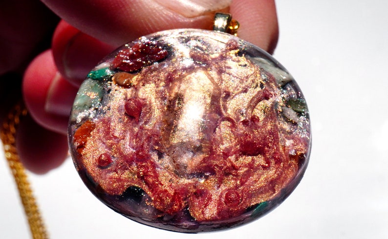 May include: Round pendant necklace with a gold chain. The pendant is made of clear resin with a swirling mix of red, gold, and green glitter.