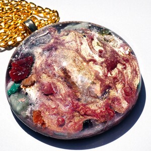 May include: A round, gold-toned pendant necklace with a clear resin inlay featuring a swirling pattern of pink, gold, and white. The resin also contains small pieces of various colored gemstones.