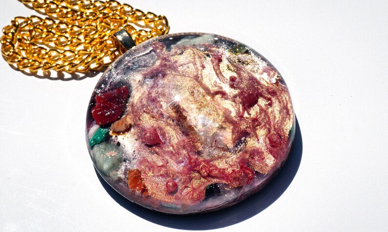 May include: A round, gold-toned pendant necklace with a clear resin inlay featuring a swirling pattern of pink, gold, and white. The resin also contains small pieces of various colored gemstones.