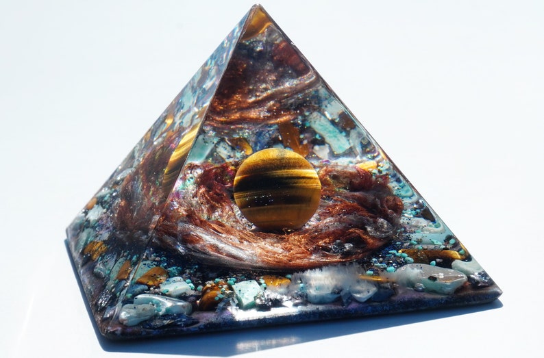 May include: A clear pyramid made of resin with a tiger's eye sphere inside. The pyramid is filled with various crystals and stones, including turquoise, gold, and copper.