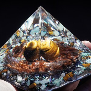 May include: A clear glass pyramid with a brown and gold tiger's eye stone inside. The pyramid is filled with small pieces of various crystals and glitter.
