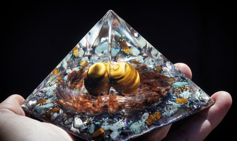 May include: A clear glass pyramid with a brown and gold tiger's eye stone inside. The pyramid is filled with small pieces of various crystals and glitter.