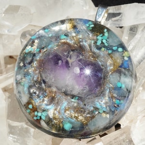 May include: A round pendant with a silver bail, featuring a central purple amethyst crystal. The pendant is encased in clear resin with turquoise, gold, and silver accents, creating a unique, colorful design. The pendant is set against a background of clear quartz crystals.
