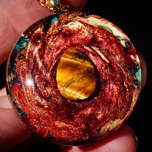 Puede incluir: Un colgante redondo con un diseño rojo y dorado en espiral. Una piedra de ojo de tigre está engastada en el centro del colgante.