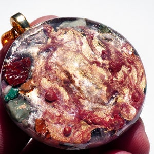 May include: A round, gold-toned pendant with a resin inlay featuring a swirling pattern of red, gold, and green. The pendant is attached to a gold chain.
