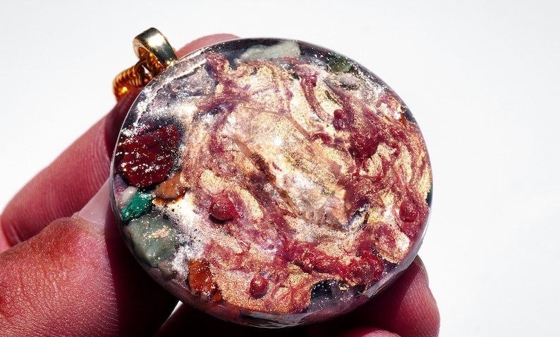 May include: A round, gold-toned pendant with a resin inlay featuring a swirling pattern of red, gold, and green. The pendant is attached to a gold chain.