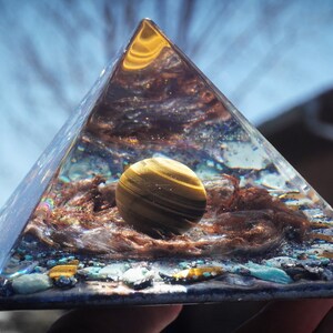 May include: A clear glass pyramid with a golden tiger's eye sphere inside. The pyramid is filled with various crystals and stones, including turquoise and copper.