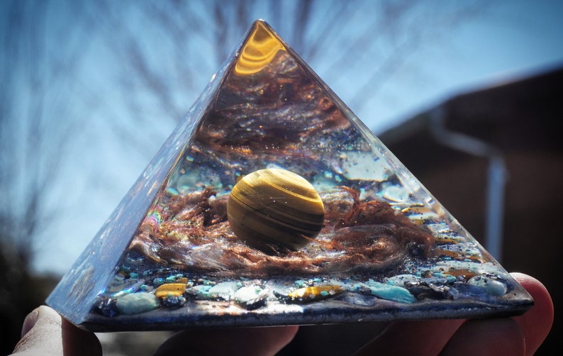 May include: A clear glass pyramid with a golden tiger's eye sphere inside. The pyramid is filled with various crystals and stones, including turquoise and copper.