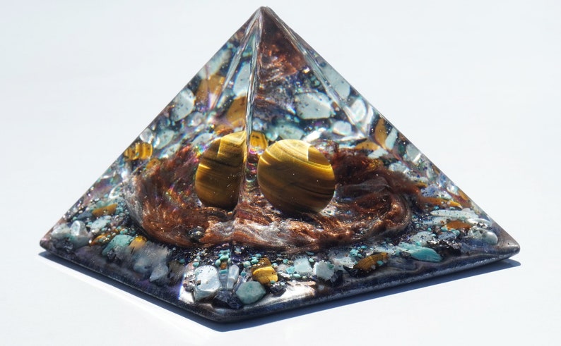 May include: A clear pyramid with a brown, gold, and blue crystal inside. The pyramid is made of resin and has a smooth, polished finish.