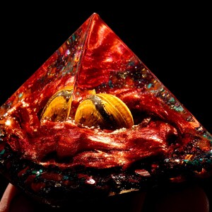 May include: A red, orange, and gold pyramid-shaped crystal with two tiger's eye stones inside. The crystal is filled with glitter and other small crystals.