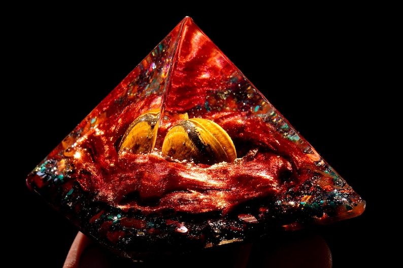 May include: A red, orange, and gold pyramid-shaped crystal with two tiger's eye stones inside. The crystal is filled with glitter and other small crystals.