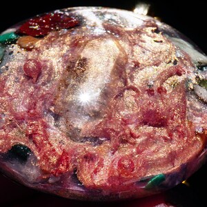 May include: A round, clear resin pendant with a pink, gold, and green swirl pattern. The pendant is filled with small pieces of glitter and other materials.