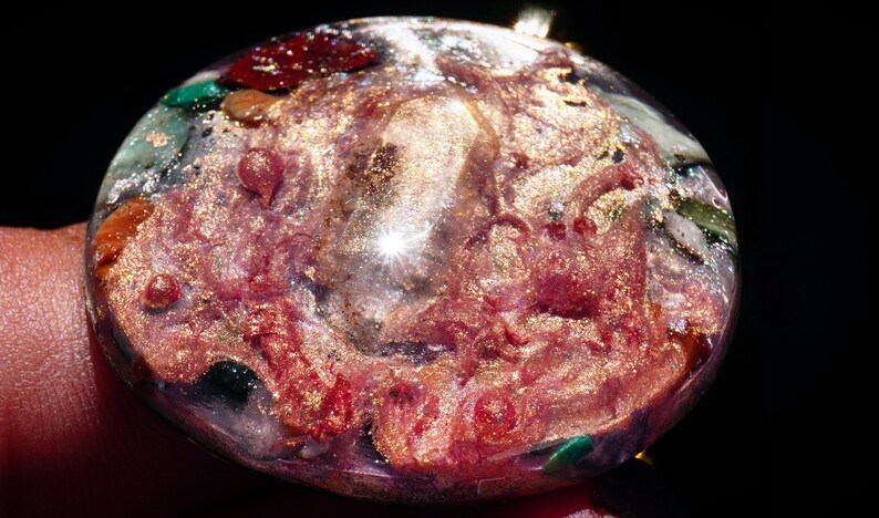 May include: A round, clear resin pendant with a pink, gold, and green swirl pattern. The pendant is filled with small pieces of glitter and other materials.