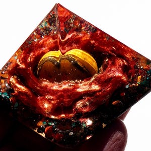May include: A red, orange, and green crystal pyramid with a yellow and gold crystal inside. The pyramid is made of clear resin and has a shiny, metallic finish.