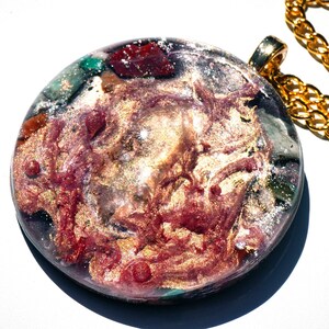 May include: A round, gold-toned pendant with a resin inlay featuring a swirling mix of red, gold, and green colors. The pendant is attached to a gold chain.