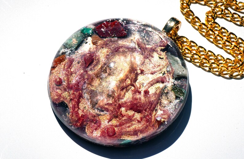 May include: A round, gold-toned pendant with a resin inlay featuring a swirling mix of red, gold, and green colors. The pendant is attached to a gold chain.