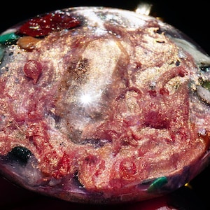 May include: A round, clear resin pendant with a pink, gold, and green swirl pattern. The pendant is filled with small pieces of glitter and other materials.