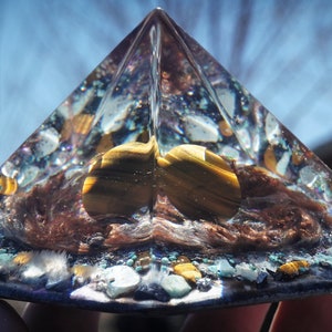 May include: A clear glass pyramid with a brown, gold, and blue interior. The pyramid is filled with small stones and crystals, including tiger's eye.