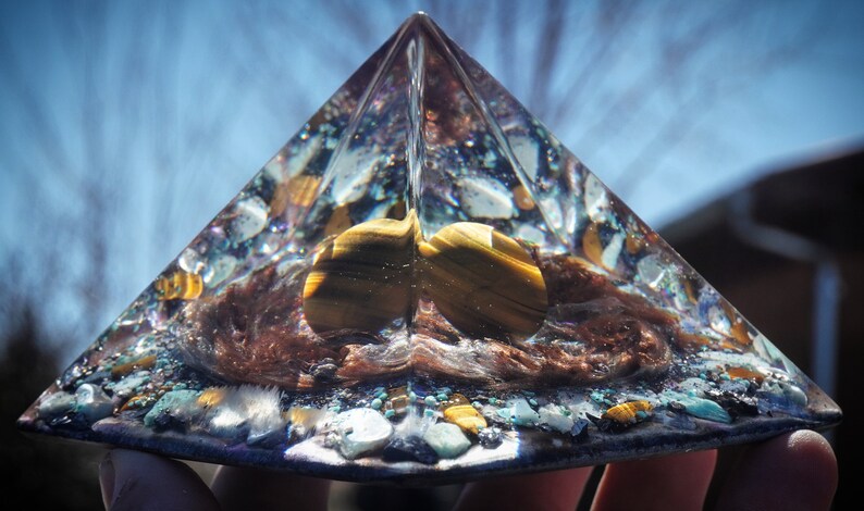 May include: A clear glass pyramid with a brown, gold, and blue interior. The pyramid is filled with small stones and crystals, including tiger's eye.