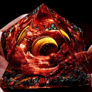 May include: A red, gold, and green pyramid-shaped crystal with a swirling pattern inside. The crystal is made of resin and has a shiny, glossy finish.